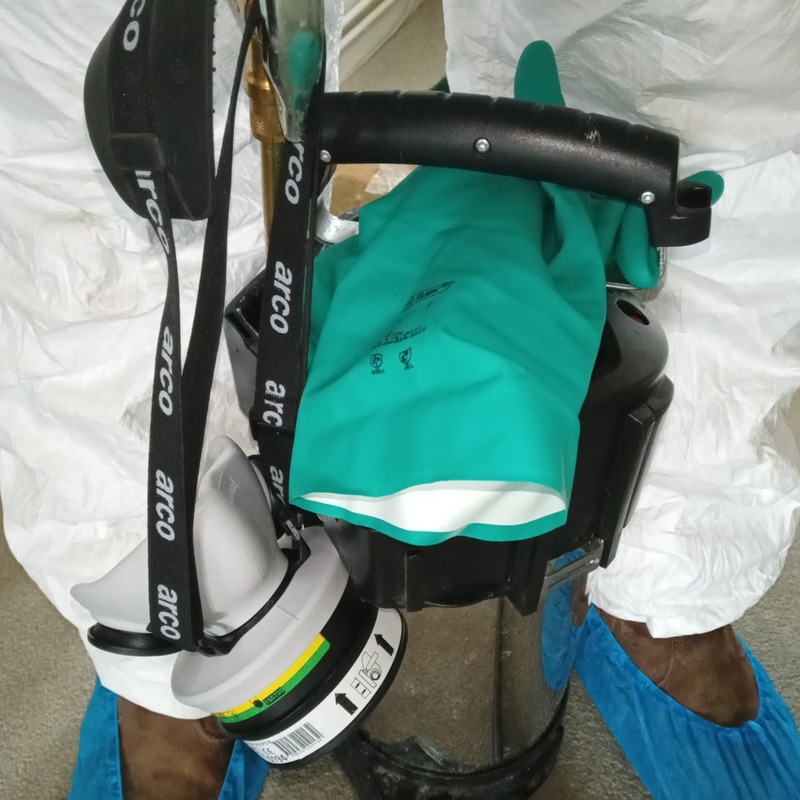 A pest controller's legs, with their feet in blue booties and a large canister in the middle. Also shown are a pair of green gloves.
